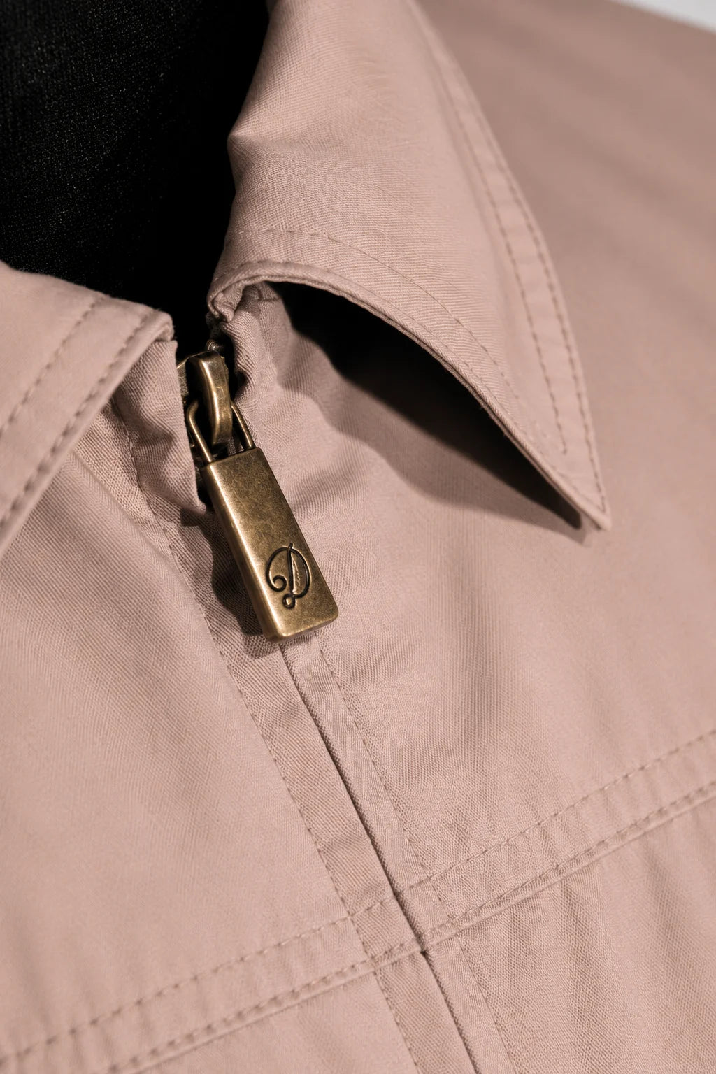 Close-up of a light beige or blush-toned jacket with a pointed collar, visible zipper, and a metallic antique-brass zipper pull engraved with a monogram logo.