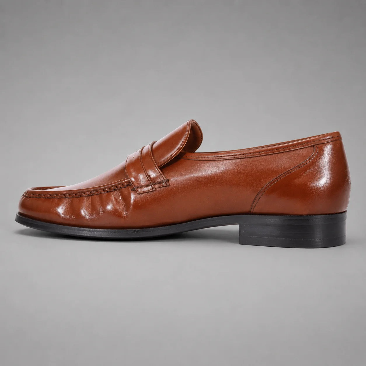 John Drake brown leather moccasin loafer with a gold-tone metal emblem on the strap, set against a neutral gray background.