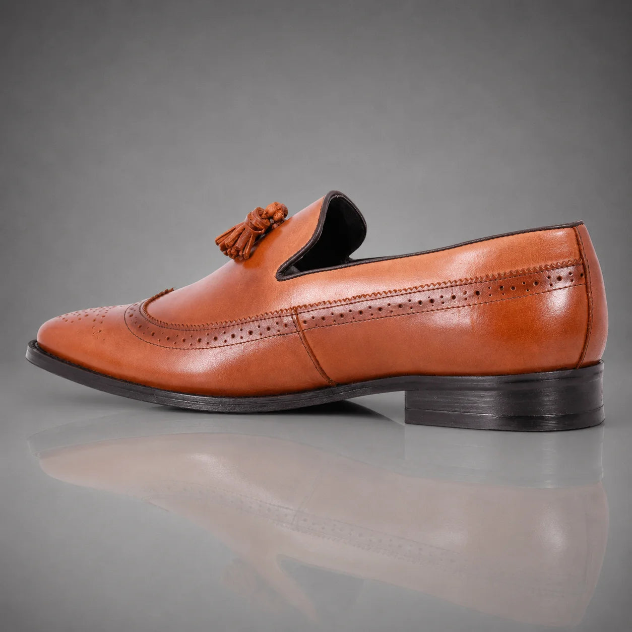 A single men's brown leather John Drake tassel loafer with brogue detailing, shown in a side profile against a gray gradient background.