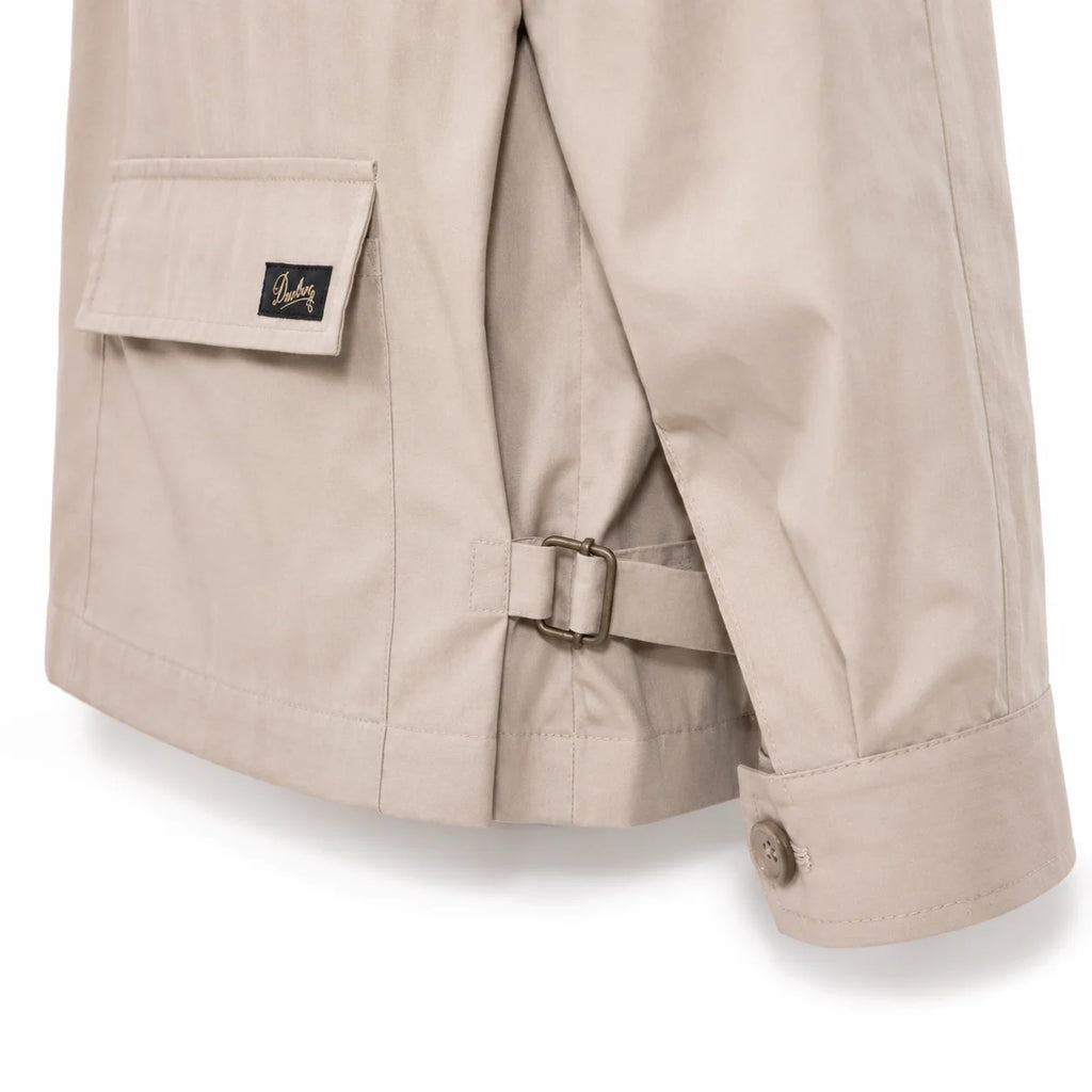 Close-up of a beige trench coat showing the back pocket with a flap and a small black logo patch, a side strap with metal buckle, and the cuff with a button.