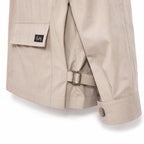 Close-up of a beige trench coat showing the back pocket with a flap and a small black logo patch, a side strap with metal buckle, and the cuff with a button.
