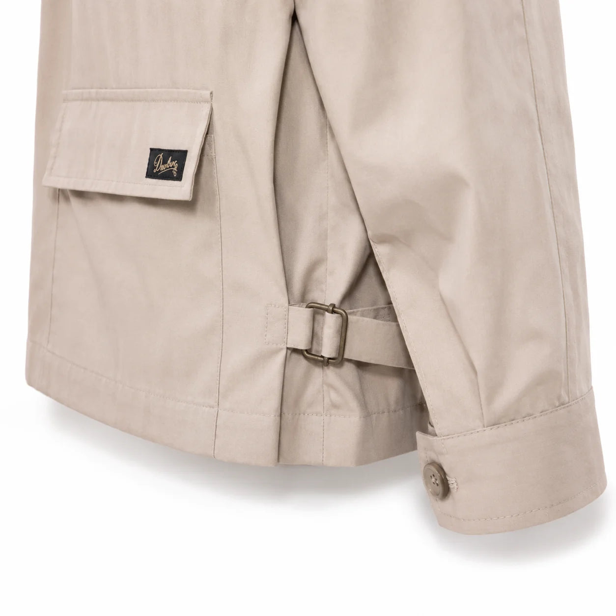 Close-up of a beige trench coat showing the back pocket with a flap and a small black logo patch, a side strap with metal buckle, and the cuff with a button.