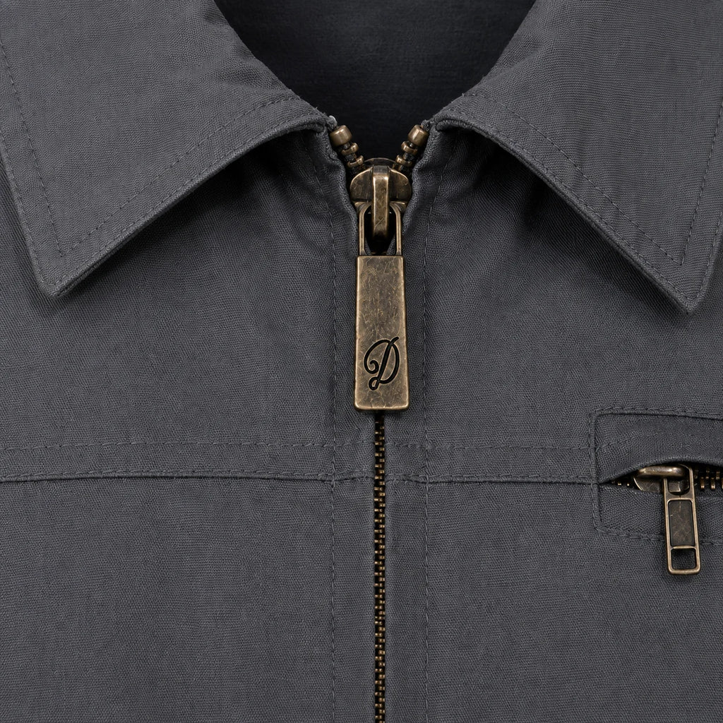 Close-up of a dark grey Durburg Mistral collared jacket with an antique bronze zipper pull featuring an engraved script 'D' logo, and a matching zippered chest pocket.