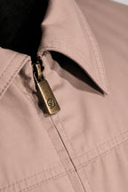 Close-up of a light beige or blush-toned jacket with a pointed collar, visible zipper, and a metallic antique-brass zipper pull engraved with a monogram logo.