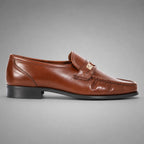 John Drake brown leather moccasin loafer with a gold-tone metal emblem on the strap, set against a neutral gray background.