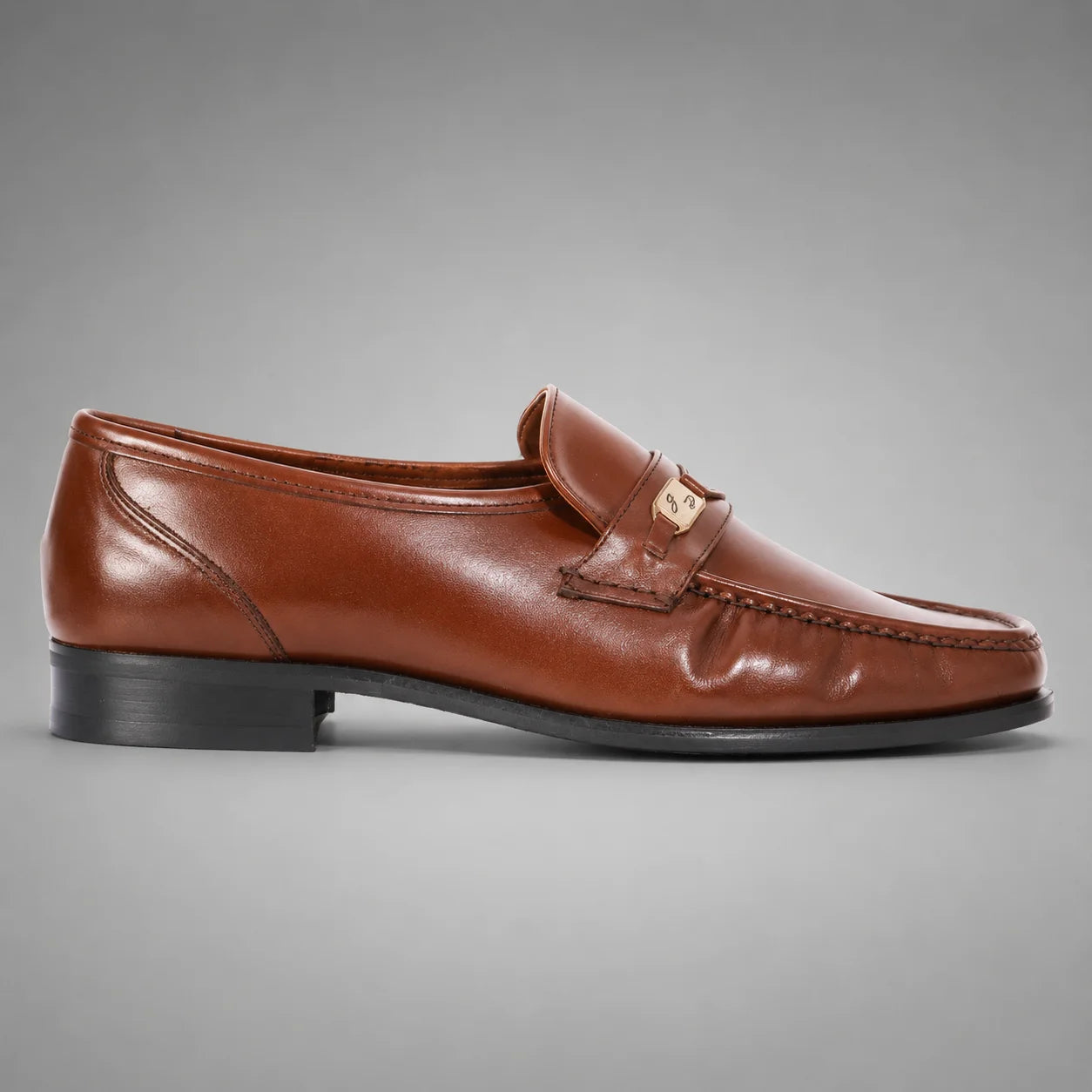 John Drake brown leather moccasin loafer with a gold-tone metal emblem on the strap, set against a neutral gray background.