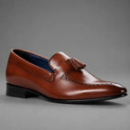 A single brown leather tassel loafer shoe with brogue detailing and a low black heel, photographed on a neutral gray background.