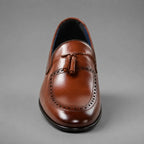 A single brown leather loafer with a tassel detail and brogue perforations, photographed from the front against a neutral gray background.