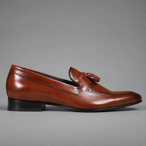 A single brown leather loafer with a tassel detail on the vamp, photographed from the side against a neutral gray background.