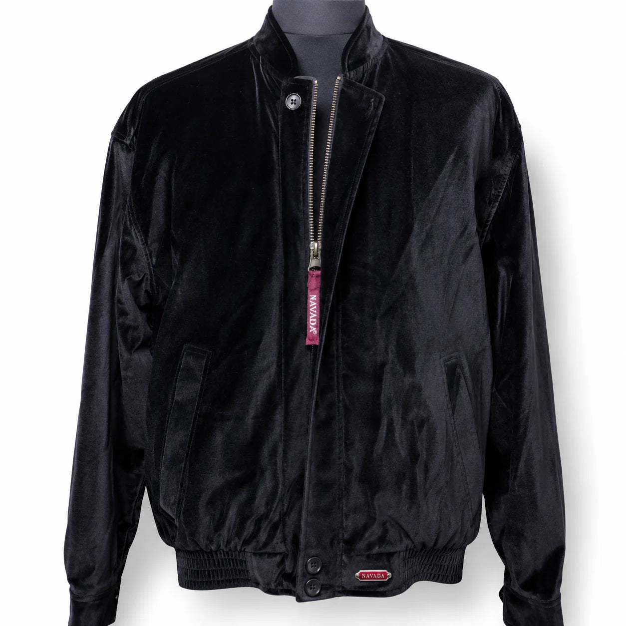 A black velvet bomber jacket displayed on a dark mannequin torso against a white background. The jacket features a silver zipper with a pink 'NAVADA' tag, side pockets, and a small metal 'NAVADA' logo badge near the bottom hem.