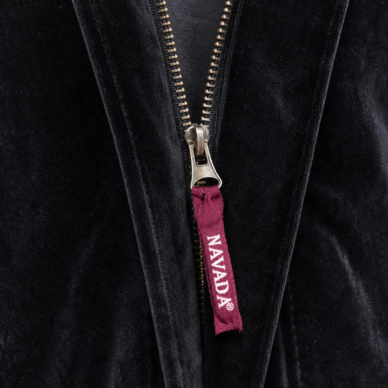 Close-up of a black velvet zippered garment with a silver zipper pull featuring a burgundy tab printed with the brand name 'NAVADA'.