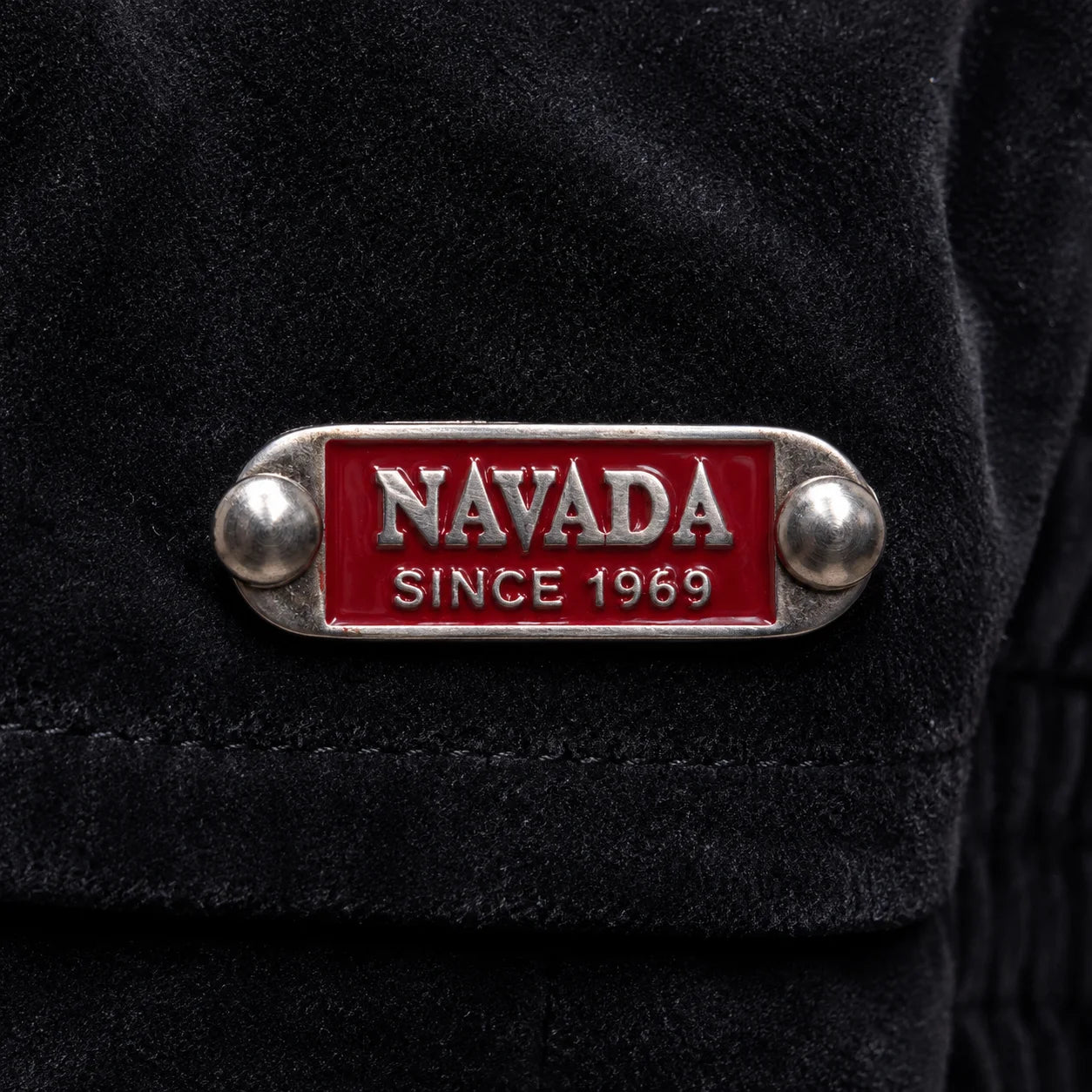 A close-up of a black textured garment featuring a silver-tone metal lapel pin with a red enamel background and the text 'NAVADA SINCE 1969'.