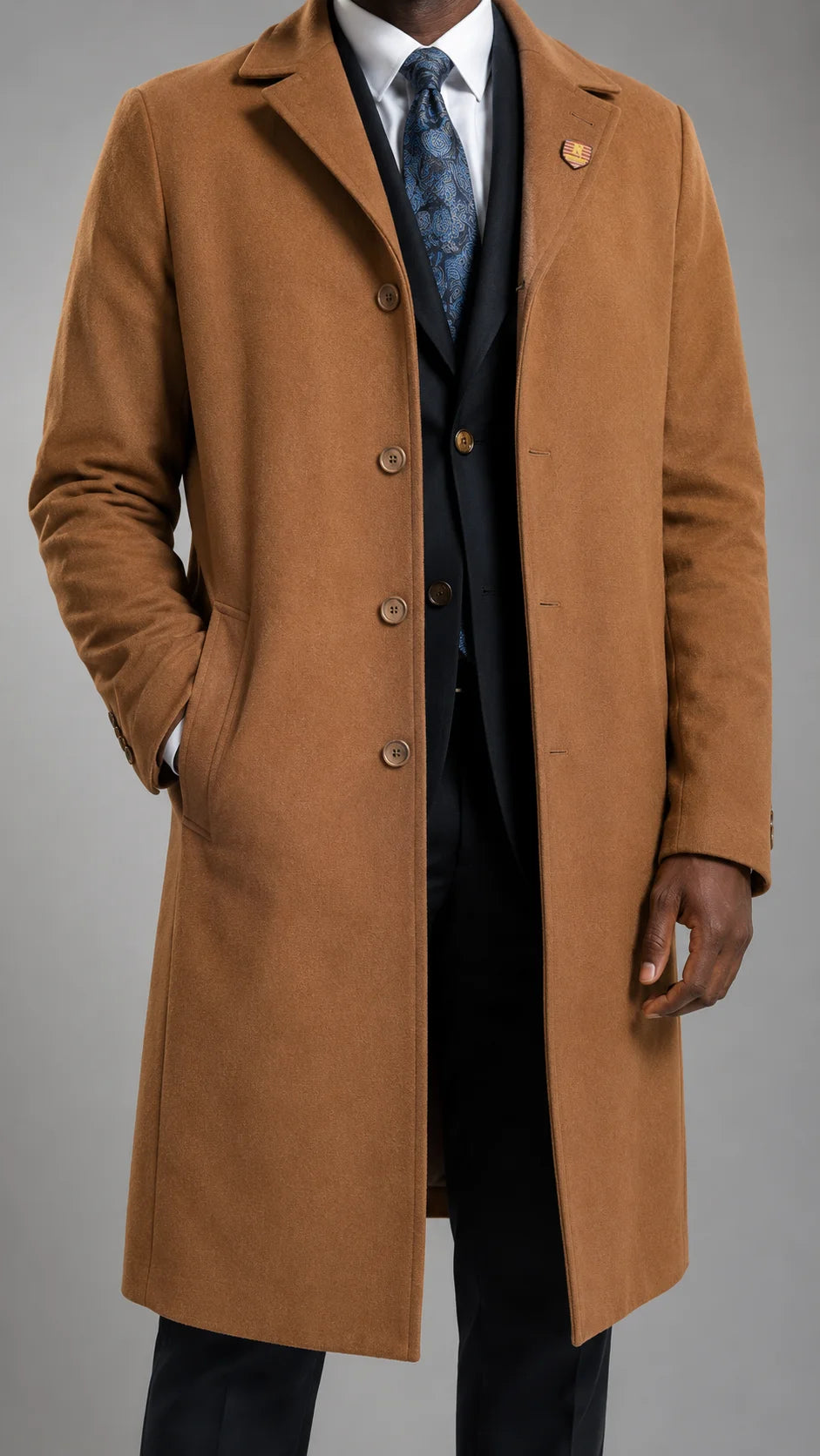 A man wearing a camel brown vashmere blend overcoat over a dark suit, white shirt, and patterned tie, photographed against a neutral gray studio background.