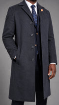 A man wearing a dark charcoal gray cashmere blend overcoat over a dark navy three-piece suit, white dress shirt, and blue patterned tie, photographed against a neutral gray studio background.