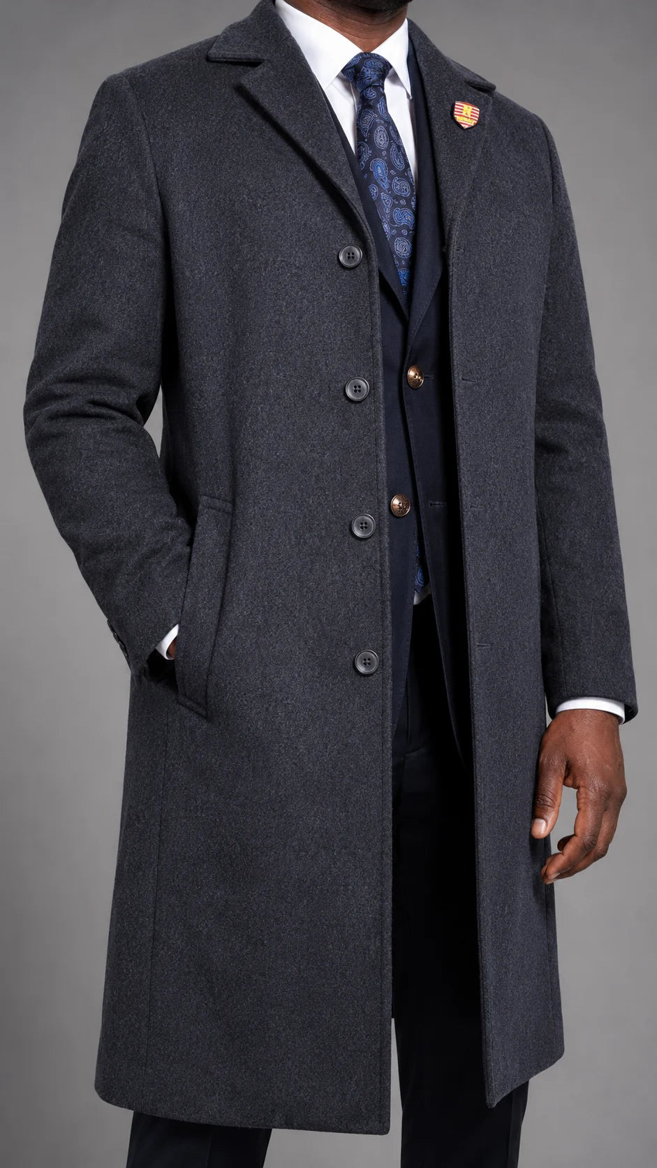 A man wearing a dark charcoal gray cashmere blend overcoat over a dark navy three-piece suit, white dress shirt, and blue patterned tie, photographed against a neutral gray studio background.
