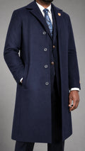 A man wearing a dark navy blue cashmere overcoat over a matching navy suit, white dress shirt, and a blue paisley tie, photographed against a neutral gray studio background.